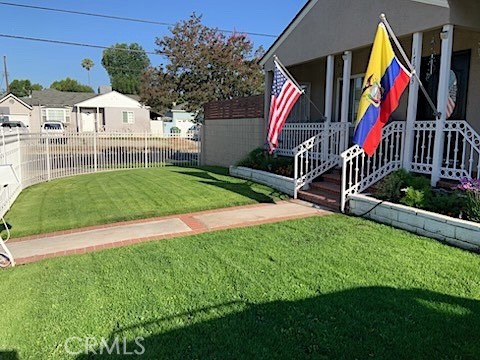 The front yard of 17513 Tiara St looking toward the covered porch, with white railed steps, a brick walkway crossing the lawn, a raised planter bed, and the fenced corner of the lot